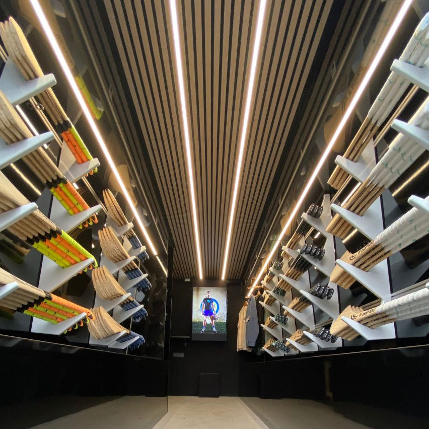 A narrow retail aisle with shelving on both sides displaying many packaged items, likely stationery or similar flat goods. The ceiling has linear LED lighting and a slatted wood panel design.